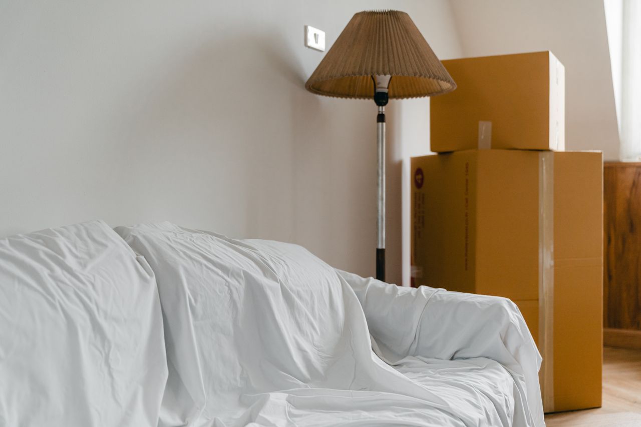 A sofa and floor lamp covered with sheets, along with packed cardboard boxes, in a bright, roomy space inside a new contemporary house